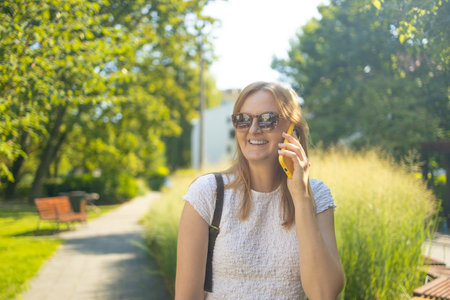 Portrait of smiling 30s student freelancer woman looking at the camera talking on phone in summer park. People urban lifestyle concept. High quality photoの写真素材