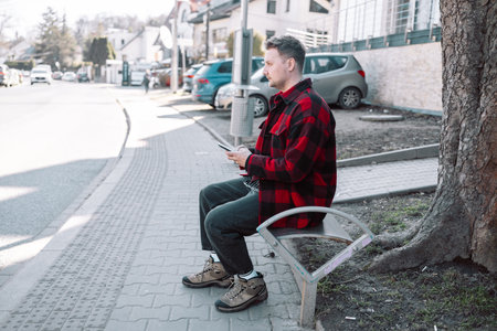 Male student with smartphone wearing plaid shirt sitting on bench while waiting for transport at tram or bus station. City public transport infrastructure, citizen and tourists transportation conceptの写真素材