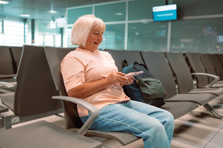 A cropped image of a senior Caucasian female tourist is holding her smartphone and passport, checking her flight information on her phone while sitting at a waiting seat in the airport terminal.の写真素材