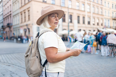 A senior woman stands in a beautiful city square, holding a map while enjoying sightseeing. She is dressed in a comfortable outfit, wearing a hat and sunglasses.の写真素材