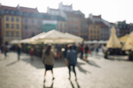 Out-of-focus image of a sunny public square with people walking near outdoor cafes in...