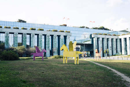 Bright yellow geometric sculpture of a horse with wings, placed on a grassy urban square. Minimalist and playful public art installation made of painted metal. Symbolic and eye-catching outdoor artwork with blue sky in the background.の写真素材