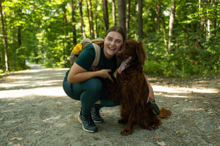 Smiling young woman in outdoor gear kneels beside her Irish Setter dog on a forest path. Their...