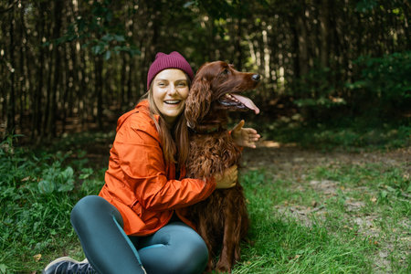 Young woman in sportswear hugging her fluffy dog in the forest while laughing. Lifestyle, friendship, nature, pet care and joy captured in one shot.の写真素材
