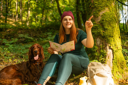 Young woman in hiking clothes sits under a tree in the forest, reading a map with her dog by her side. A backpack and reusable water bottle lie nearby. Peaceful nature break during an outdoor adventure.の写真素材