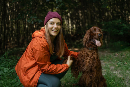 A cheerful young woman wearing an orange jacket and beanie poses with her Irish Setter dog in a forest setting. The pair is surrounded by greenery and natural light filtering through the trees.の写真素材