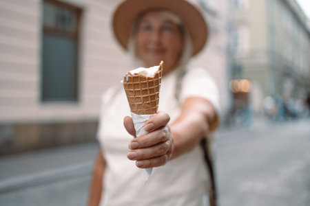 Cheerful senior woman holding a cone of ice cream on a pleasant day in the city, enjoying her treat while strolling down the street. A perfect moment of relaxation and indulgence.の写真素材
