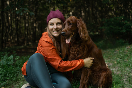 Smiling woman sitting beside her Irish Setter in a forest, enjoying peaceful moments in nature. Great for themes of pet companionship and outdoor leisure.の写真素材