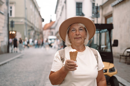 60s senior woman walking leisurely down a city street with a cone of ice cream, enjoying a moment of calm and happiness under the sun. A perfect blend of relaxation and indulgence.の写真素材
