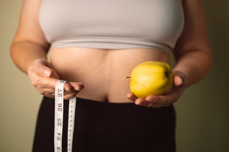 A woman is holding a yellow apple and a measuring tape around her waist, symbolizing health, weight loss, and body care. She wears a gray sports bra, showcasing self-care and wellnessの写真素材