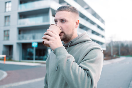 Handsome young man drinking coffee. Man with beard hand holding paper coffee cup going walk outside on city streetの写真素材