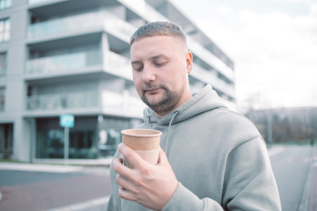 Handsome young man drinking coffee. Man with beard hand holding paper coffee cup going walk outside on city streetの写真素材