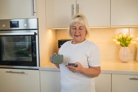 A senior woman stands in her kitchen holding a phone and a coffee cup, smiling. This image highlights the modern life of seniors enjoying their daily routines with technology and comfortの写真素材