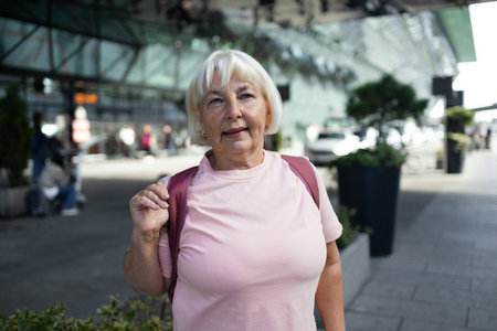 Happy elderly woman in a pink t-shirt with a backpack, smiling at the airport terminal. She ready for her next travel adventure. Ideal for travel, senior lifestyle, and vacation content.の写真素材