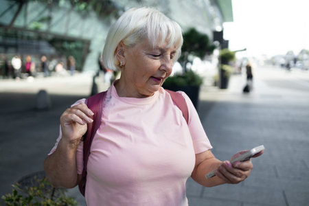 An elderly woman is standing outside the airport terminal, smiling while checking her smartphone. She appears to be receiving or sending a message, with her backpack resting on her shoulder.の写真素材