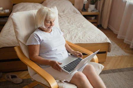 Elderly woman sits in a cushioned chair using a laptop in a softly lit bedroom. The setting highlights comfort, digital literacy, remote communication, and modern lifestyle for older adults.の写真素材