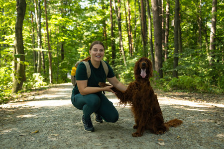 Smiling young woman in outdoor gear kneels beside her Irish Setter dog on a forest path. Their joyful interaction highlights companionship, nature bonding, and the spirit of adventure in the wild.の写真素材