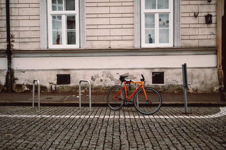 Bright orange bicycle is parked and secured to a metal bike rack on a cobblestone street. Behind it stands a historic beige building with white-framed windows, evoking a calm, urban European settingの写真素材