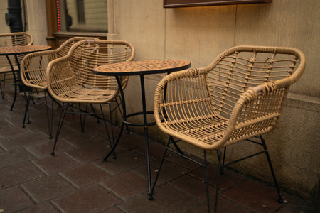 Cozy outdoor cafe setup featuring wicker chairs and round tables on a wet cobblestone street. The inviting scene captures a charming spot ideal for casual conversation or coffee breaks in an urban settingの写真素材