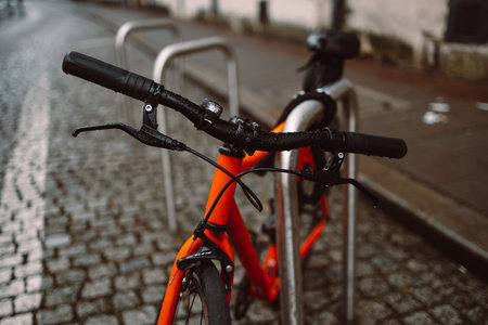 Close-up of an orange bicycles handlebars, locked to a metal bike rack on a cobblestone street. The wet pavement and blurred background evoke a quiet, rainy day in the city.の写真素材
