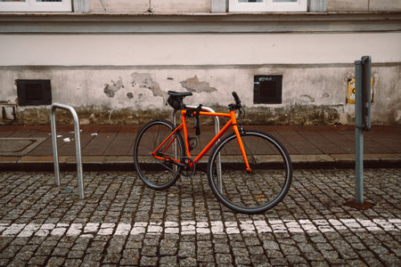 Bright orange bicycle is parked and secured to a metal bike rack on a cobblestone street. Behind it stands a historic beige building with white-framed windows, evoking a calm, urban European settingの写真素材