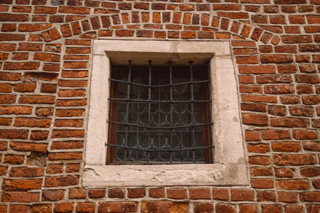 A small, square window with iron bars set into an aged red brick wall. Framed by light stone, the window evokes a historical or fortified architecture style, adding texture and character to the structure.の写真素材