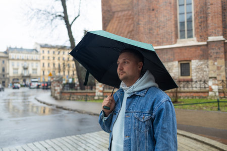 Man in a denim jacket and hoodie walks through a rainy European street, holding a large black umbrella. The background features historic architecture and wet cobblestones, capturing a moody, overcast day.の写真素材