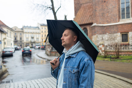 Man in a denim jacket and hoodie walks through a rainy European street, holding a large black umbrella. The background features historic architecture and wet cobblestones, capturing a moody, overcast day.の写真素材