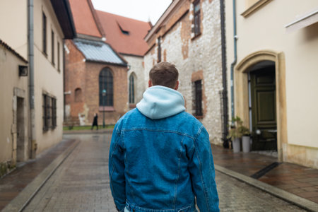 A man in a denim jacket with a hood walks down a quiet, cobbled street on a rainy day. The surrounding buildings are historic, with stone walls and arched windows, evoking a calm European atmosphere.の写真素材