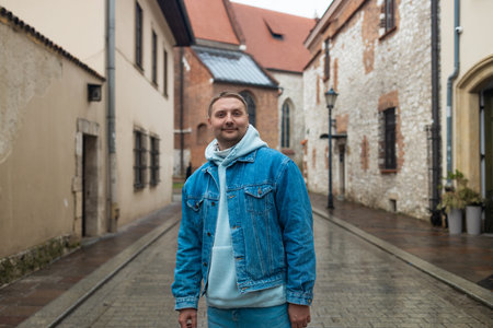 A casually dressed man in a denim jacket and hoodie smiles while standing on a cobbled street in a historic European town. The rainy weather and old stone buildings create a charming, peaceful mood.の写真素材