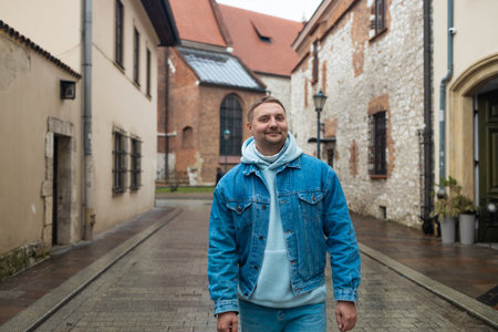 A casually dressed man in a denim jacket and hoodie smiles while standing on a cobbled street in a historic European town. The rainy weather and old stone buildings create a charming, peaceful mood.の写真素材