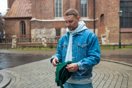 Man in a denim jacket and hoodie is seen folding a green umbrella on a cobblestone street, with a historic brick building in the background. The wet pavement hints at recent rain, creating a peaceful, reflective atmosphere.の写真素材