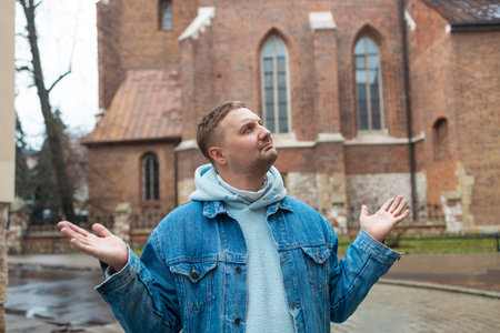 Casually dressed man in a denim jacket and hoodie stands on a cobblestone street with a puzzled or indifferent expression, shrugging his shoulders in front of a historic brick building. A light drizzle hints at moody weather.の写真素材