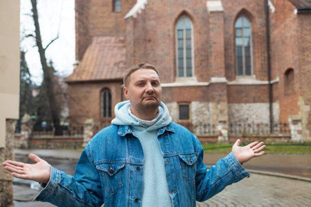 A man in a denim jacket and hoodie stands in front of an old brick church, shrugging with raised...