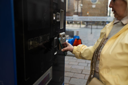 A woman in a yellow jacket uses a ticket machine in Krakow to buy a tram or bus ticket. The machine...