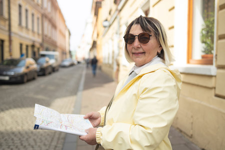 Smiling elderly woman in a yellow jacket and sunglasses holding a city map and pointing at something on a cobblestone street in a European town. She appears to be exploring or navigating her way.の写真素材
