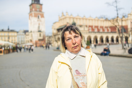 An older woman stands in Krakow Main Market Square, with the iconic Cloth Hall and Town Hall Tower visible in the background. The sunny day highlights the charm of the historical city center.の写真素材