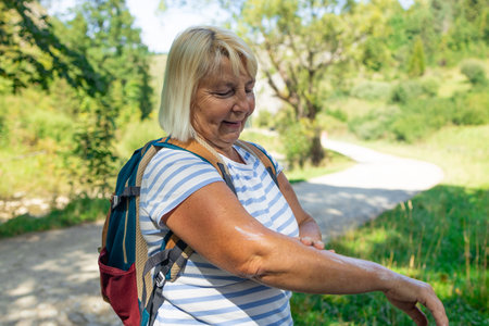 Smiling senior woman with a backpack applying cream or insect repellent on her arm during a summer hike. Captured in a scenic forest setting, this moment reflects self-care, outdoor lifestyle, and active aging.の写真素材