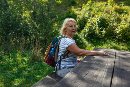 Elderly female hiker relaxing at a wooden picnic table in the forest. Wearing a backpack and enjoying the peaceful atmosphere during her outdoor nature walk.の写真素材