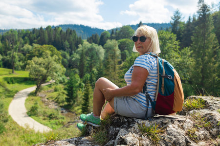 Active senior woman with a backpack and red cap sitting peacefully on a rocky cliff overlooking a lush green forest. She enjoys a scenic view during a summer hike, with bright sunlight and blue skies above.の写真素材