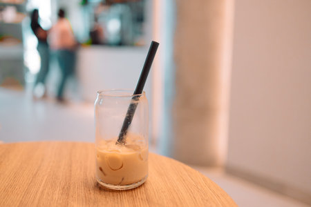 Partially finished glass of iced coffee with a black straw on a wooden table inside a bright modern...