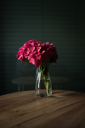 Elegant bouquet of pink hydrangea flowers in transparent glass vase placed on wooden table with dark background. Concept of still life, home decoration, and natural beauty. Minimalist floral arrangement symbolizing elegance, romance, interior design, and peaceful atmosphere in modern lifestyle and home decor.の写真素材
