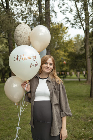 Smiling pregnant woman standing in park holding festive balloons with inscriptions Mommy to be and...