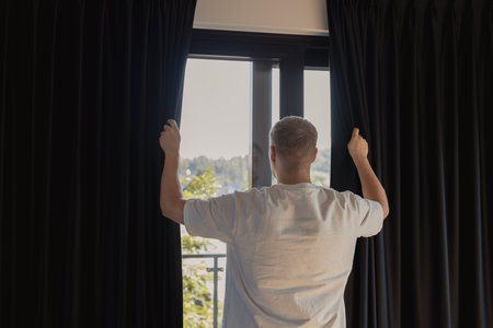 Back view of man in white clothes opening dark curtains on window in modern home interior. Concept of morning routine, lifestyle, and daily wellness. Symbol of new day, light, awakening, and comfort in household life, connected with relaxation, positivity, and healthy living.の写真素材