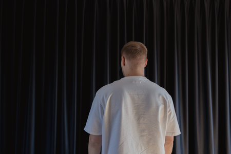Back view of man in white clothes opening dark curtains on window in modern home interior. Concept...