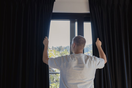 Back view of man in white clothes opening dark curtains on window in modern home interior. Concept of morning routine, lifestyle, and daily wellness. Symbol of new day, light, awakening, and comfort in household life, connected with relaxation, positivity, and healthy living.の写真素材