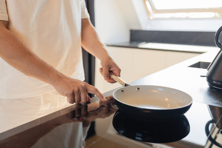 Close-up of man in white clothes putting frying pan on electric stove in modern kitchen. Concept of...