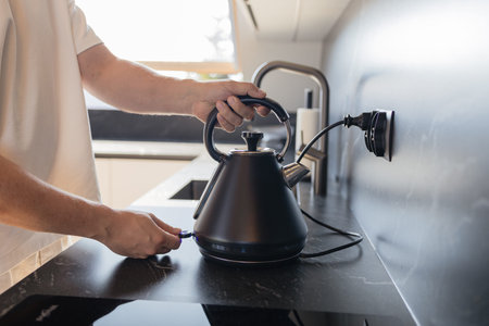 Close-up of man putting black electric kettle on stove in modern kitchen with marble wall. Concept...