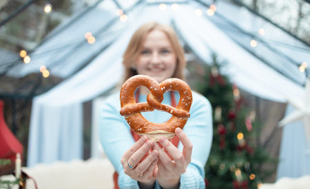 Smiling blonde Caucasian woman in a light blue sweater holding a big pretzel toward the camera. Cozy Christmas background with decorated tree, lights, and holiday atmosphere creating a warm winter mood.の写真素材
