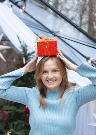 Joyful blonde Caucasian woman in a light blue sweater holding a red Christmas gift box near her face...